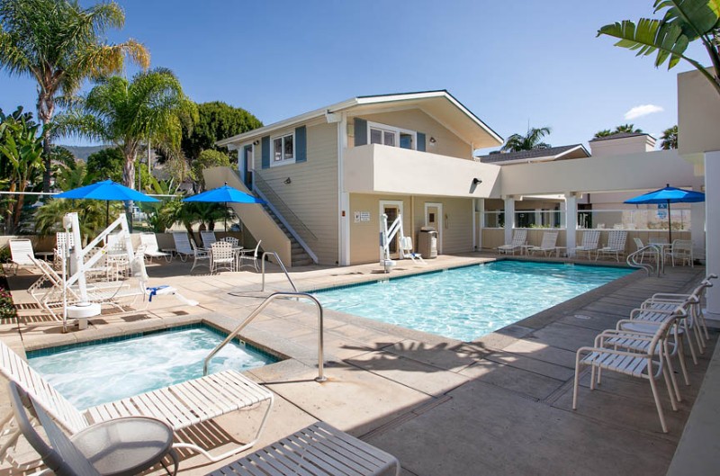 A hotel pool area with a rectangular pool, lounge chairs, and umbrellas, surrounded by a building and palm trees under a clear blue sky.