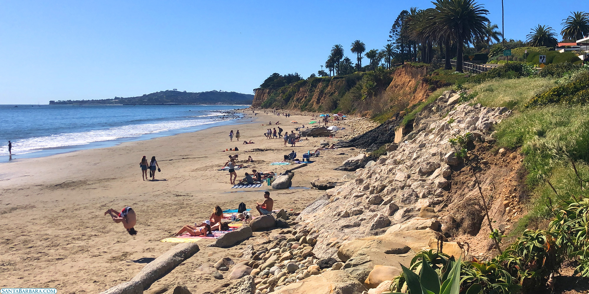 A sandy beach with people on a sunny day, featuring palm trees and rocky cliffs.